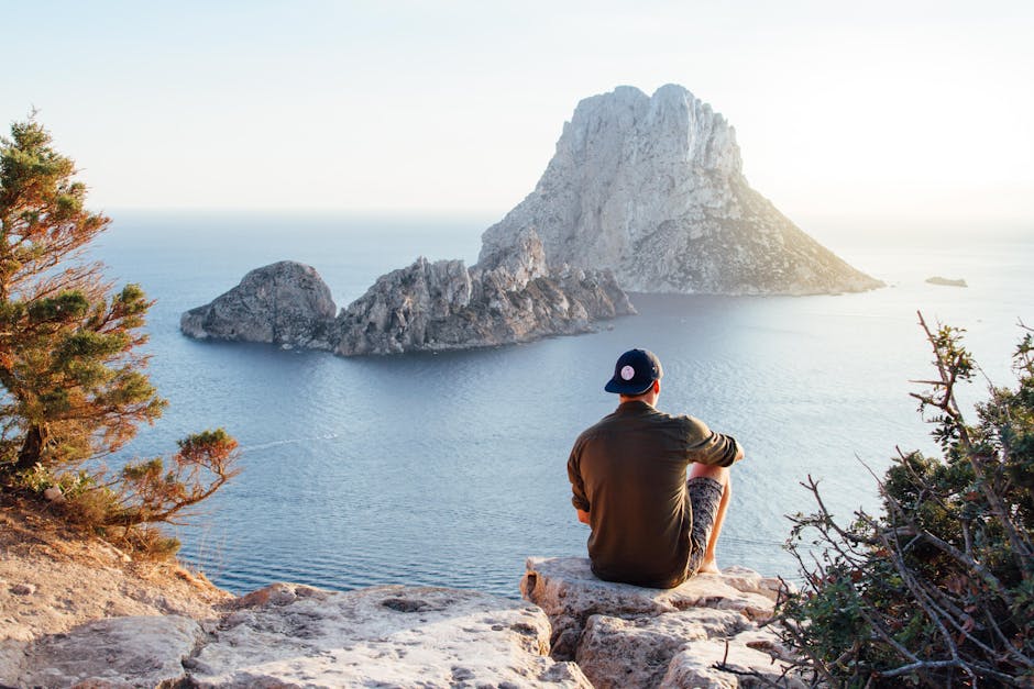 Man enjoys a scenic view of Es Vedrà at sunset from a cliff in San Juan Bautista, providing a perfect summer escape